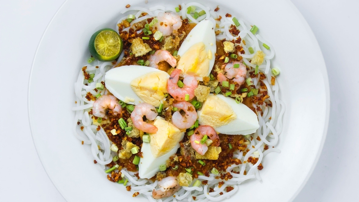 白い皿に盛り付けられた、フィリピン料理のパンシット・パラボック。白い麺の上にオレンジ色のソースがかかり、ゆで卵、小エビ、刻んだネギ、揚げた豚の皮、カラマンシーが彩り豊かにトッピングされています