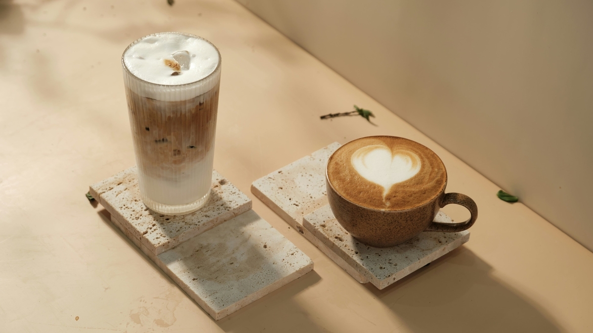 ベージュの台の上に置かれた2種類のコーヒー。左側にはミルクの層が美しいアイスカフェラテの背の高いグラス、右側にはハートのラテアートが施された温かいカフェラテのカップが、石のコースターに載っています