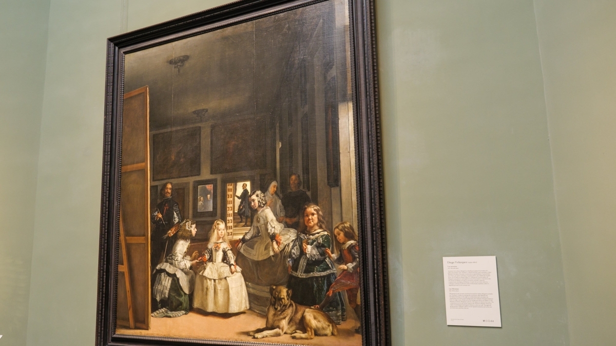 美術館の壁に展示されたベラスケスの名画『ラス・メニーナス』。黒い額縁に収められた大きなキャンバスには、幼い王女や侍女、画家本人が描かれています。右下の壁には作品解説の白いパネルが添えられています。
