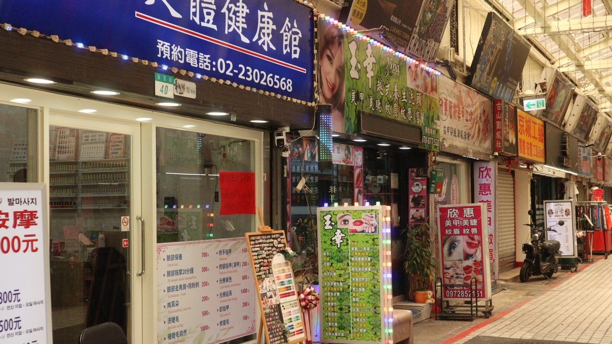 漢字の看板が並ぶ賑やかな夜の商店街。青い大きな看板には電話番号が記され、店先にはメニューが書かれたイーゼルや電飾のついた看板が立っています。タイル敷きの道を奥へと続く街並みが写っています