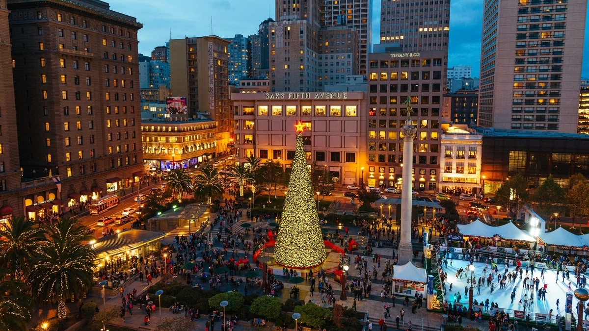 サンフランシスコのユニオンスクエアの夕景。巨大なクリスマスツリーと、周囲に広がる大勢の人々、アイススケートリンク、そしてライトアップされた街並みが写っています。