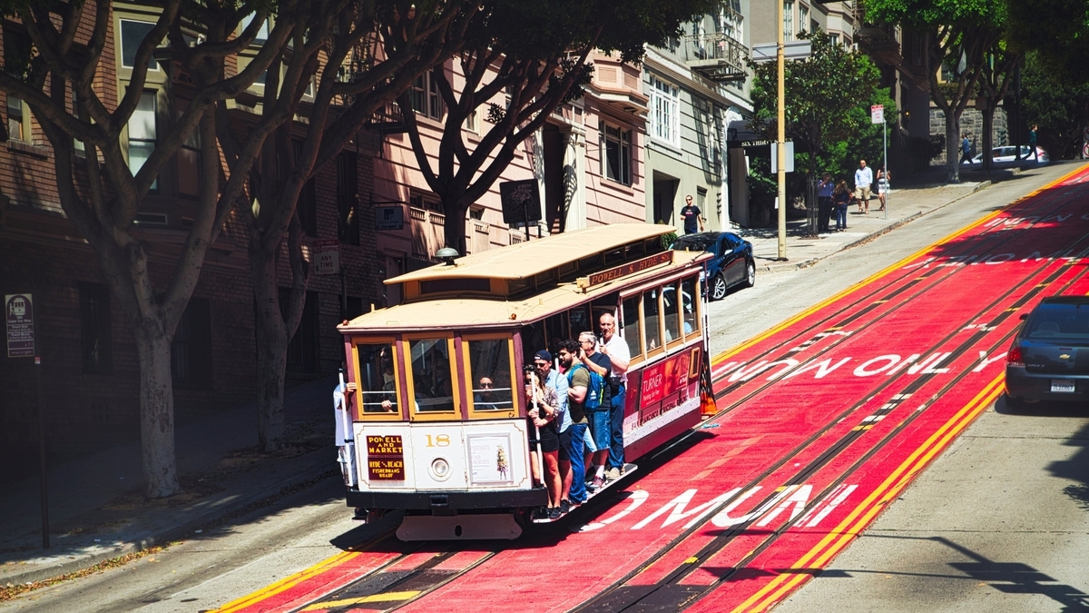 サンフランシスコの急勾配の坂道を下るケーブルカー。赤い路面には「MUNI ONLY」と白文字で書かれ、多くの乗客が乗車しています。