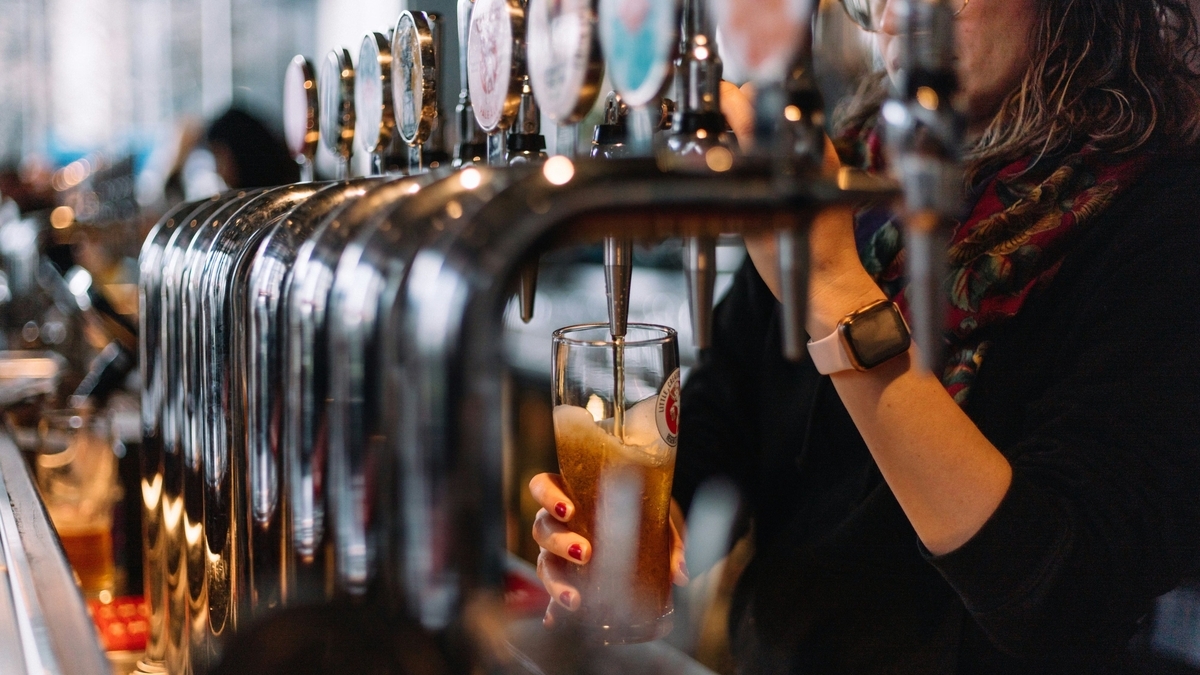 パブのカウンターで店員がビールサーバーからグラスにビールを注ぐ様子。手元にはスマートウォッチを着用しており、背景には一列に並んだタップハンドルが見えます。