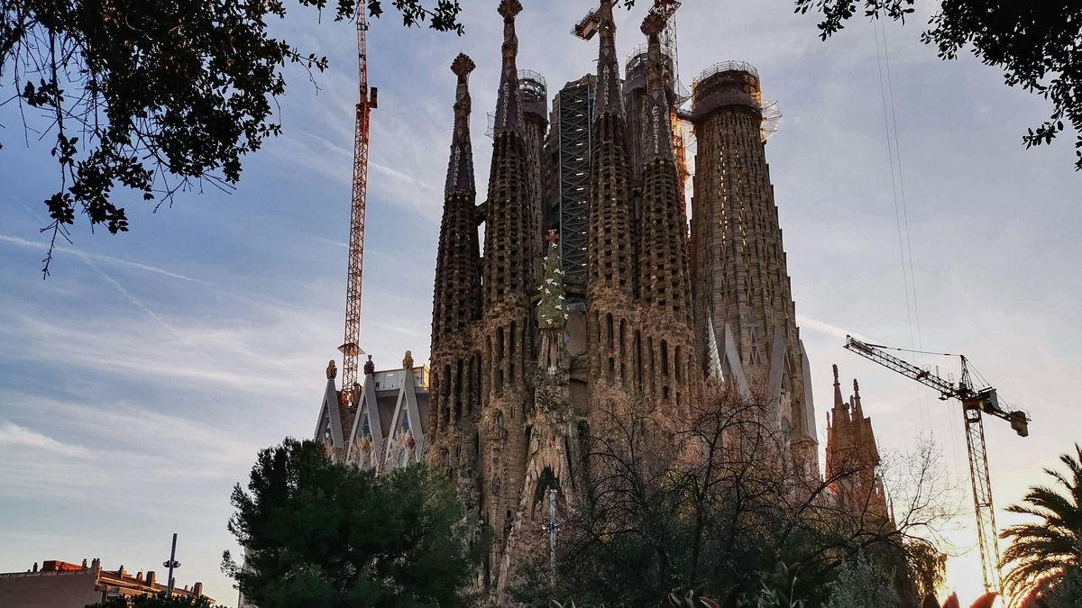 夕暮れ時の空を背景にしたサグラダ・ファミリアの外観。複雑な装飾が施された高い塔が複数立ち並び、建設途中のクレーンや手前の木々のシルエットが重なっている