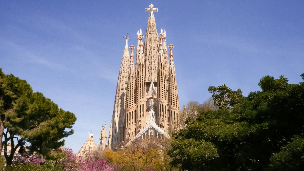 青空の下、豊かな緑の木々の合間から見えるサグラダ・ファミリアの全景。中央にそびえる高い塔の先端には十字架があり、細部まで細かな彫刻が施された茶褐色の外壁が特徴的。