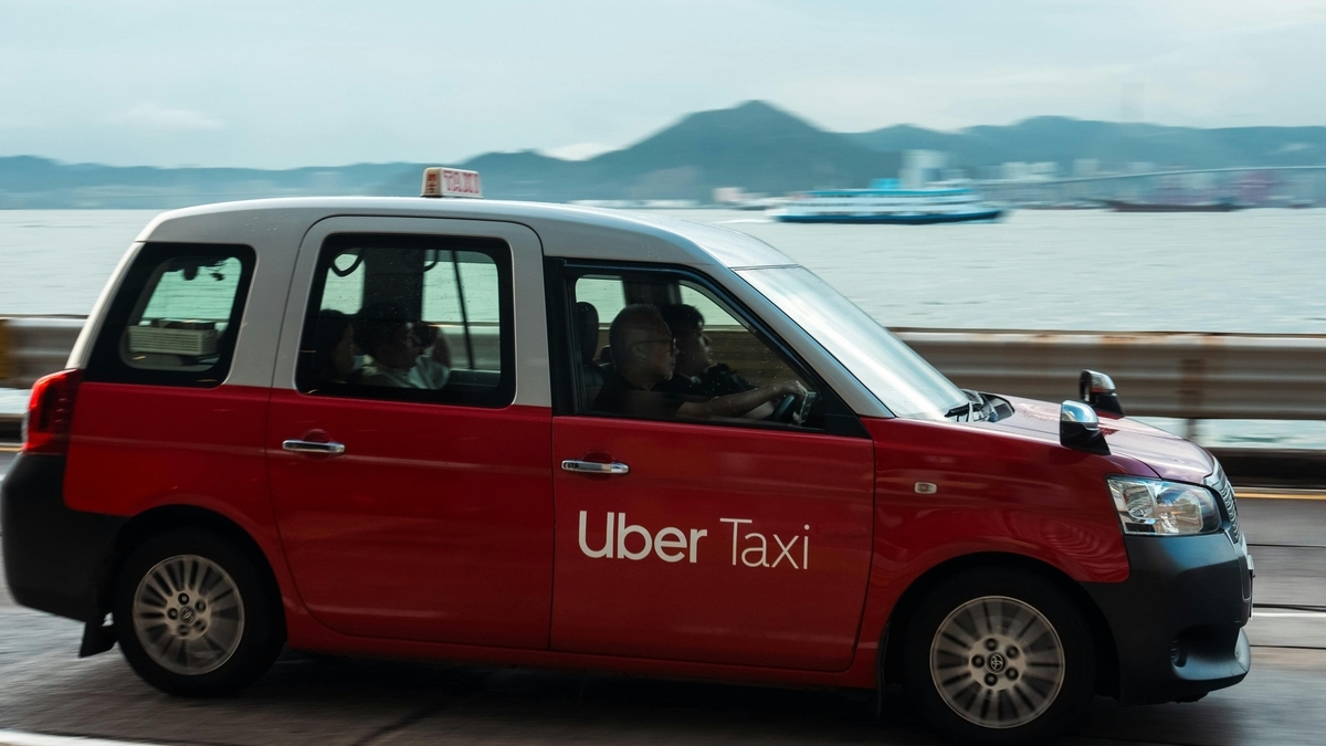 赤いボディに「Uber Taxi」のロゴが入り、屋根に「TAXI」表示があるミニバン型タクシーが、海の近くの道を走行中。車内には乗客2名と運転手が乗っています。背景は穏やかな海と山並み。