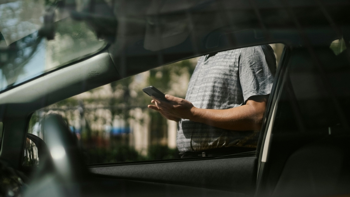 車外に立つ男性がスマートフォンを両手で操作している様子を、車の助手席側の窓越しに撮影。男性はグレーの半袖シャツを着用し、窓には外の風景と車内の影が映り込んでいます