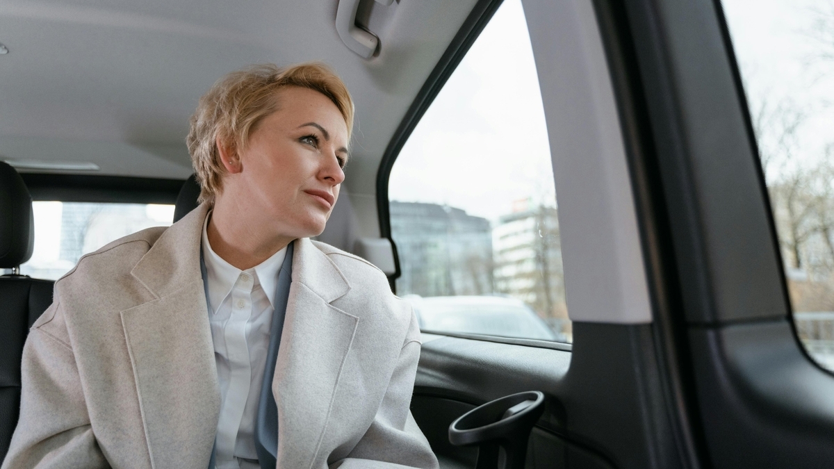車の後部座席に座る、金髪でベージュのコートを着用した女性。白シャツと水色のスカーフを合わせ、視線は車の右側にある窓から外に向けられています