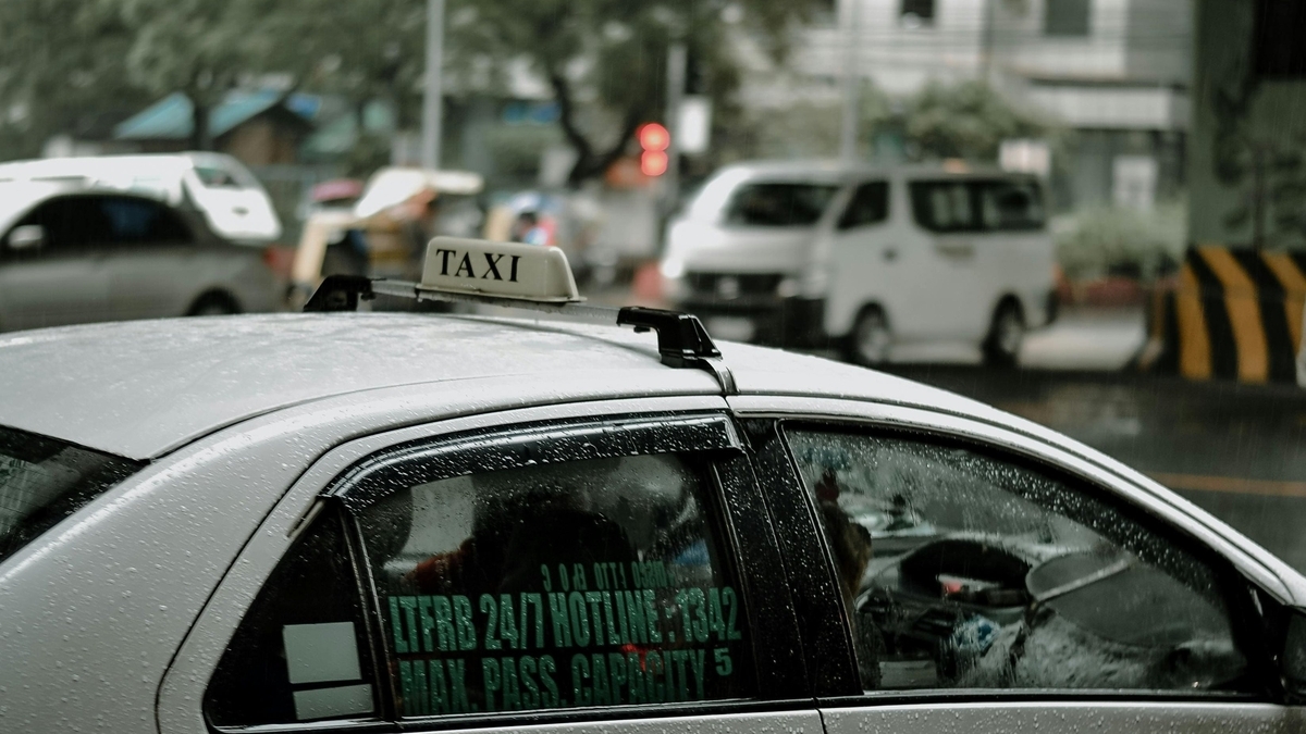 雨に濡れる白タクシーの後部座席。窓ガラスには「LTFRB 24/7 HOTLINE 1342」「MAX. PASS. CAPACITY 5」の緑文字のステッカーが貼られ、車内と前方の交通が透けて見えます