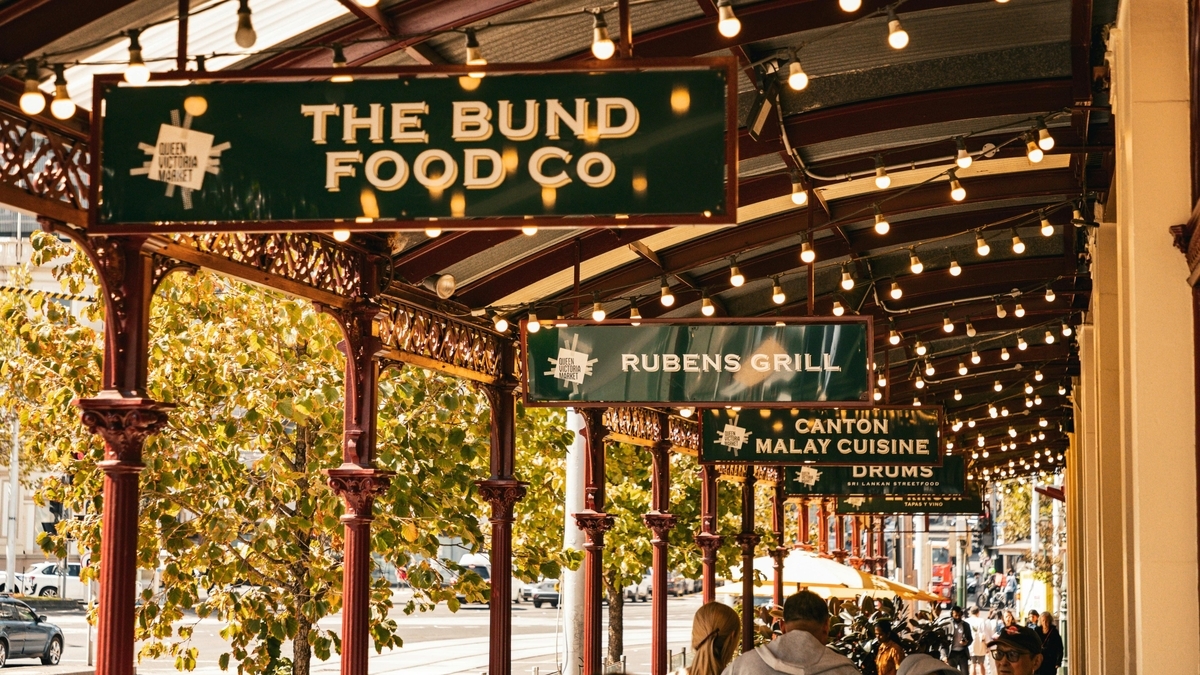 「THE BUND FOOD CO」「RUBENS GRILL」「CANTON MALAY CUISINE」といった看板が複数ぶら下がる、レトロな雰囲気のアーケード。装飾された柱や電球が連なり、背景には街路樹と車、そして歩く人々がいる。