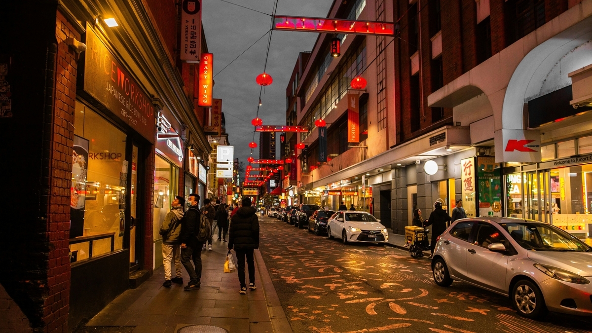 夜、ネオンサインや赤い提灯が点灯する商店街。左側に「RICE WORKSHOP」の看板を掲げた赤いレンガ造りの店舗があり、その前を数人の通行人が歩く。車道には白い乗用車とシルバーのコンパクトカーが停車しており、路面には模様が描かれている