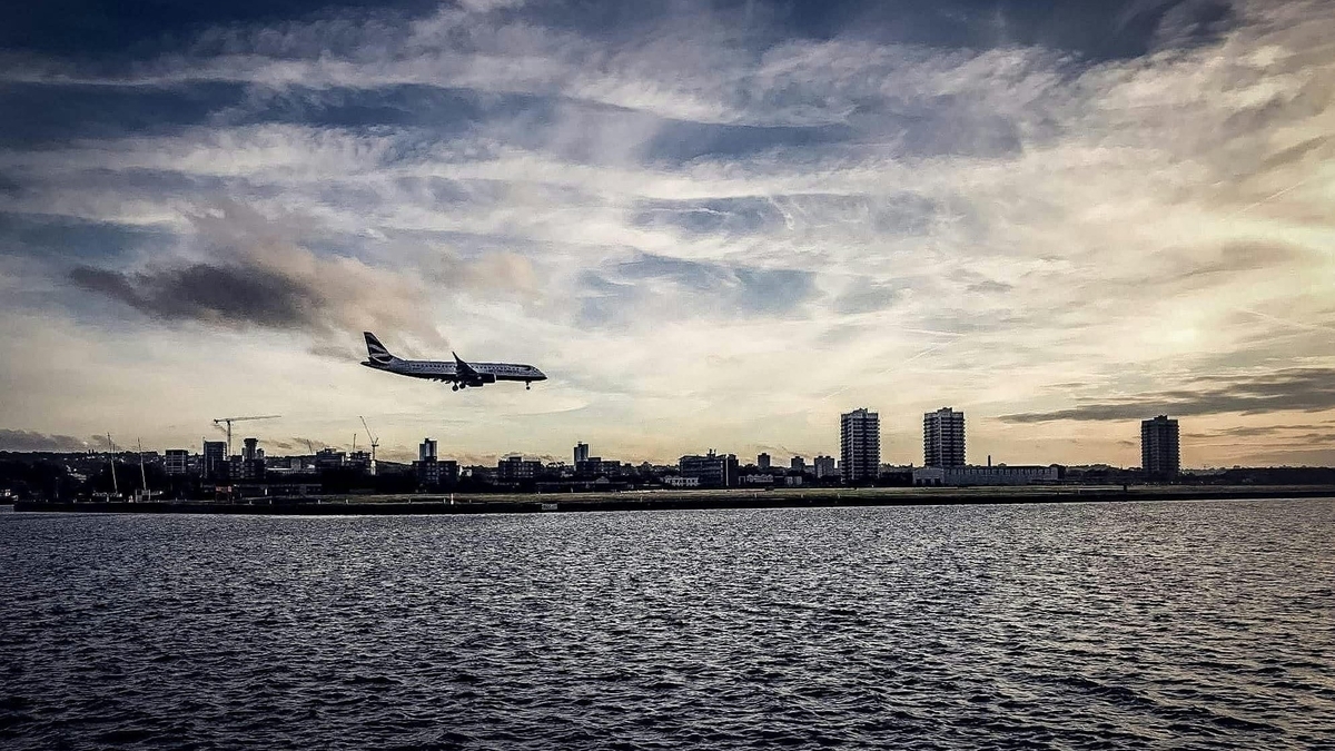 夕暮れ時の広い水辺の向こう側に、低く立ち並ぶ街の建物。空には薄い雲が広がり、一機の旅客機が低空を飛行している。手前の水面には細かな波が立ち、全体的に落ち着いた色調で構成されている。