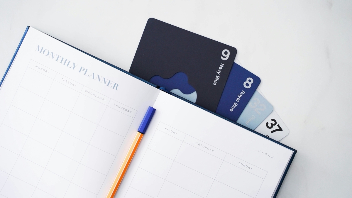 大理石調のテーブルに広げられた、MONTHLY PLANNERと書かれた手帳。横にはオレンジ色のペンが置かれ、手帳のポケットからは「Navy Blue」などの色名と番号が記されたカードが複数枚のぞいている