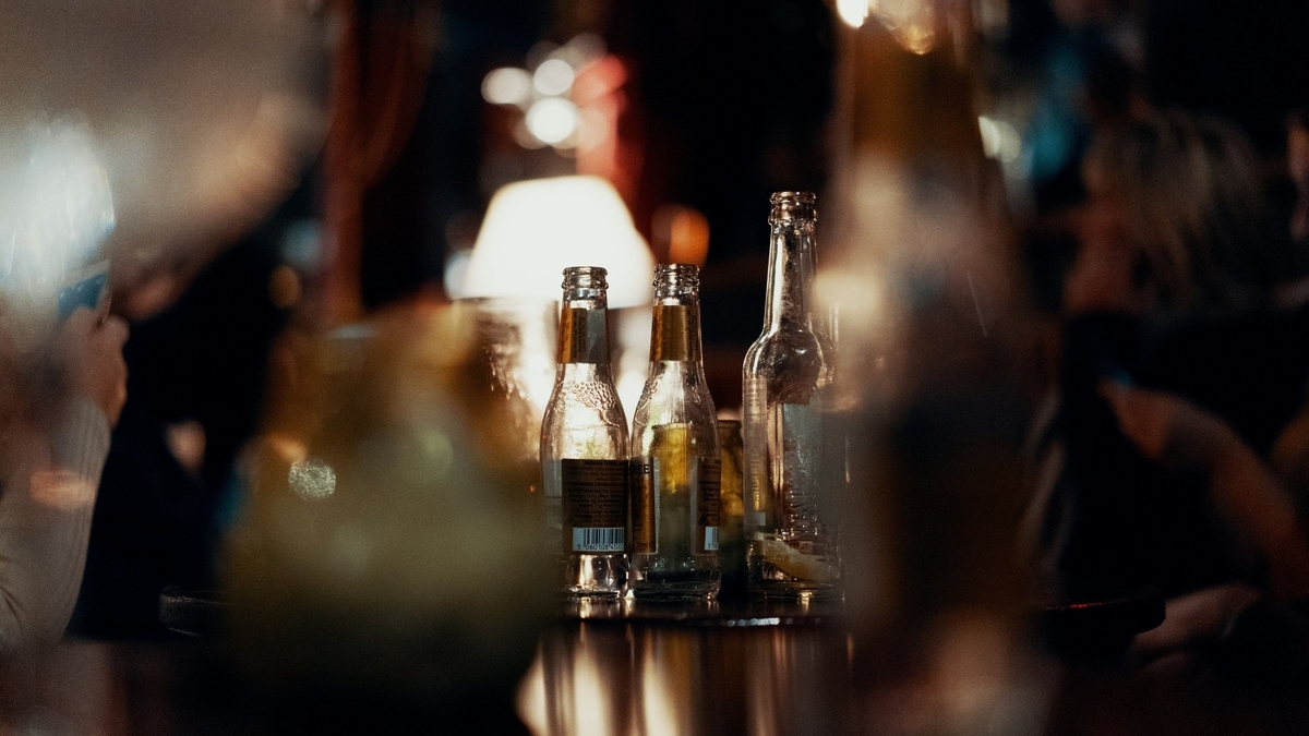 薄暗い店内のテーブルに置かれた、飲みかけのビール瓶が数本。背景には温かい色のランプや人の影がぼやけて写り、リラックスした夜の飲食店の情景を切り取っている