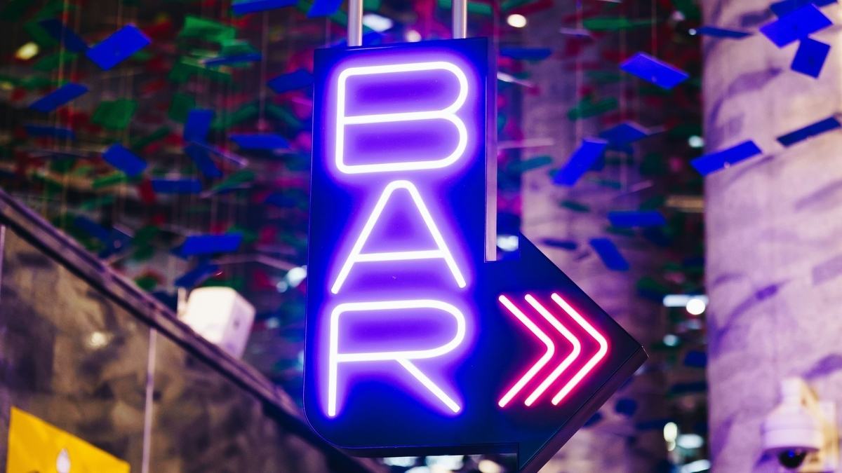 紫色のネオンで「BAR」と書かれた下向き矢印型の看板。背後には青や緑の短冊状の装飾が吊り下げられ、都会的で夜の活気を感じさせる飲食店入り口の様子