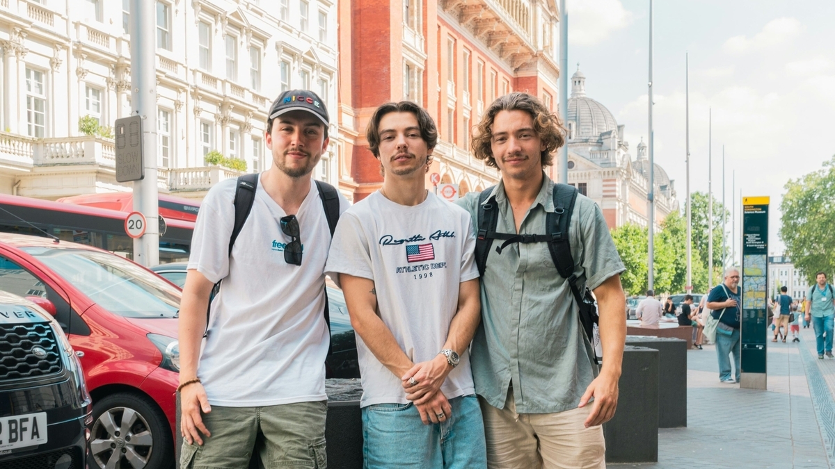 ロンドンの街並みをバックに、Tシャツやシャツを着た若い男性3人が肩を並べて立っている様子