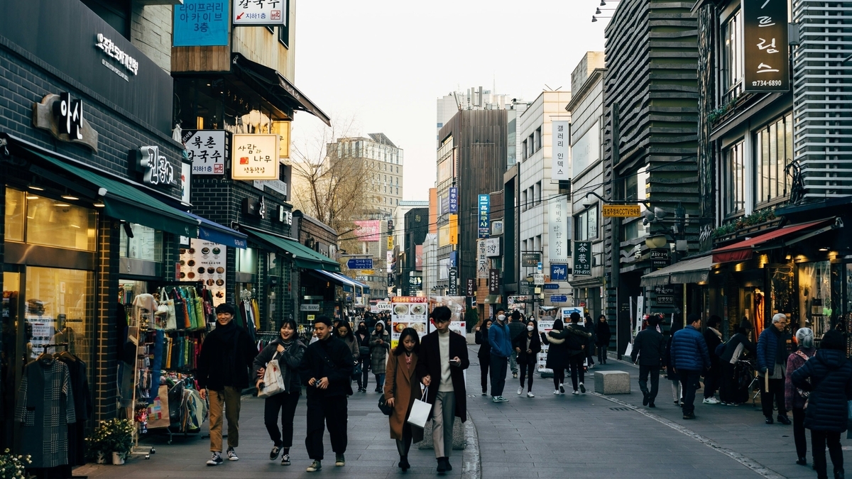 韓国語の看板が並ぶ賑やかな商店街の通りを歩く人々。建物には青や黄色の看板が掲げられ、通りの両側には衣料品店などの店舗が軒を連ねています。冬服を着た多くの観光客や地元の人々が行き交う日常の光景です