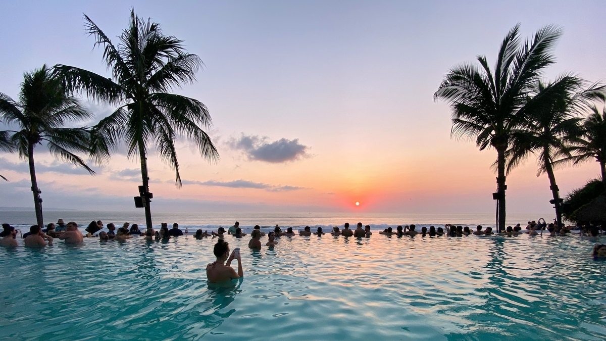 夕暮れ時のインフィニティプールで、水平線に沈む太陽を眺める多くの人々。プールの先には海が広がり、左右には背の高いヤシの木のシルエットが見えます。オレンジ色に染まった空と水面が幻想的な雰囲気を作っています