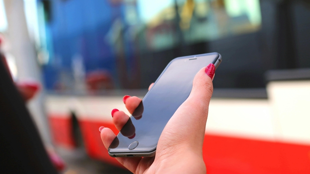 赤いネイルを施した右手がスマートフォンの黒い画面を支え、背景に赤と白の路面電車が停車している様子