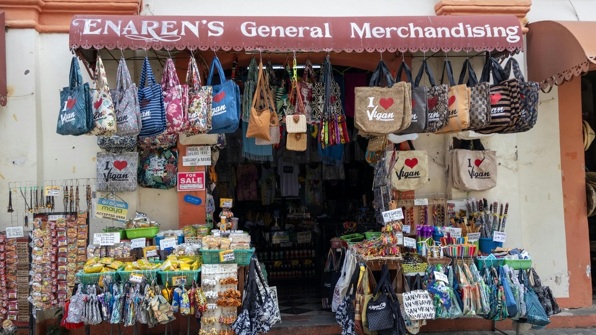 「Vigan」のロゴが入ったトートバッグや色とりどりの布製バッグが、店頭に所狭しと吊り下げられています。下部の棚にはスナック菓子や小物類が並び、赤字の看板が掲げられたフィリピンの土産物店の店先です