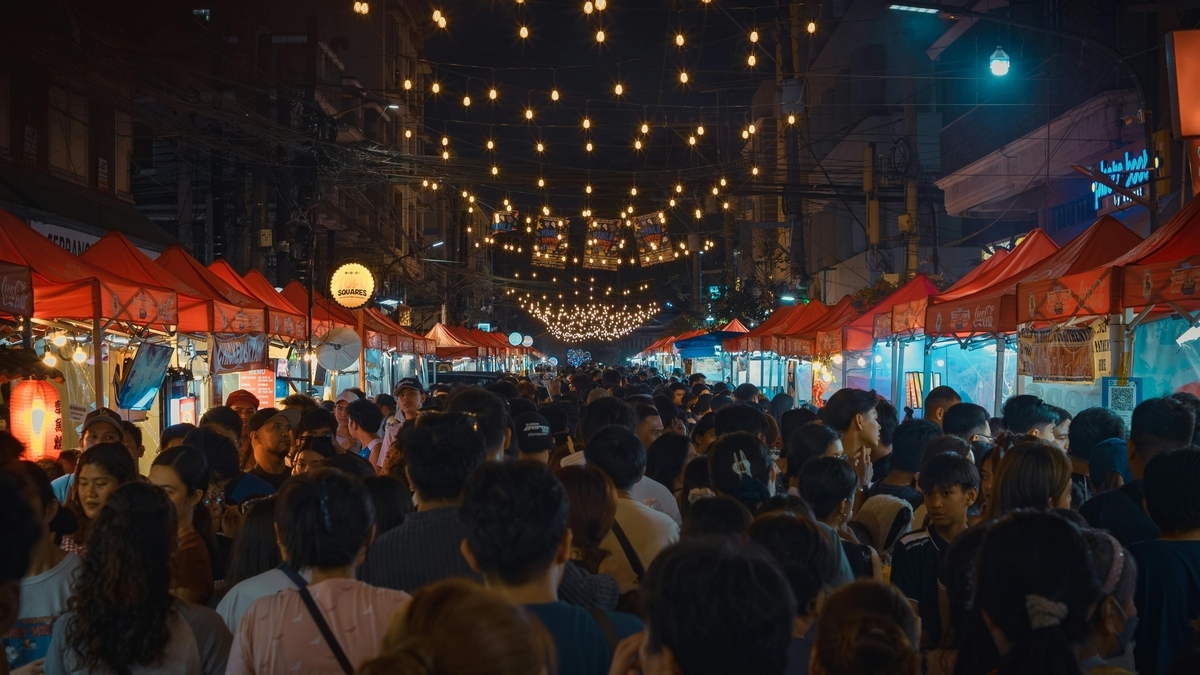 夜の街にオレンジ色の電球が連なり、大勢の人々で賑わう通りです。両脇には赤い屋根の屋台が並び、看板や電飾の明かりが人々の顔を照らしています。奥まで続く光の列が活気ある夜市の光景を作り出しています