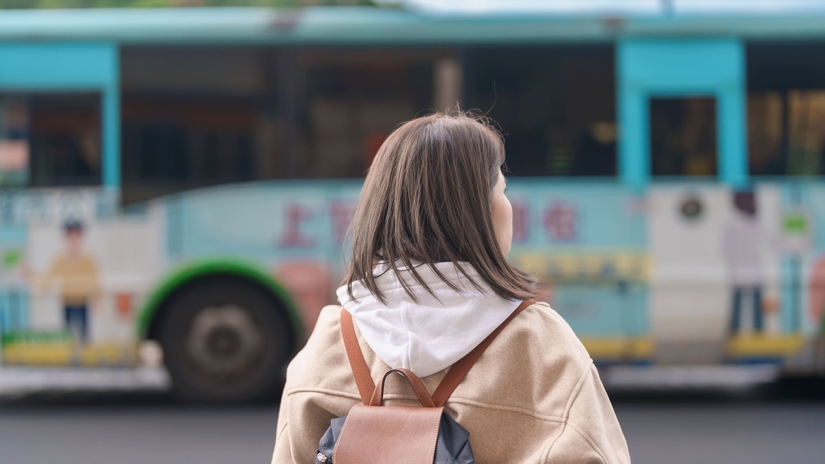 ベージュのコートとリュックを背負った女性が、水色の路線バスが停車する道路を見つめている後ろ姿。バスの車体には文字が書かれており、背景は街中の風景が少しぼやけて写っている。