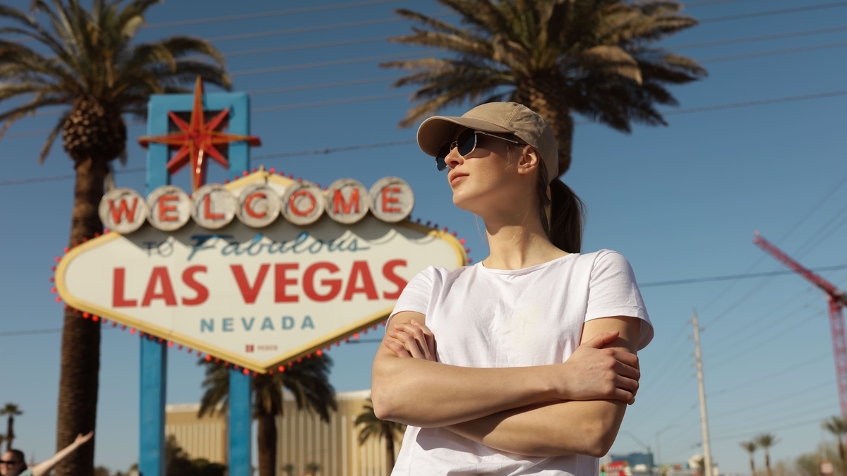 「WELCOME TO Fabulous LAS VEGAS」の看板の前に立つ、白いTシャツとキャップ姿の女性。腕を組み、視線を斜め上に向けている。背景には青空と背の高いヤシの木が写っている