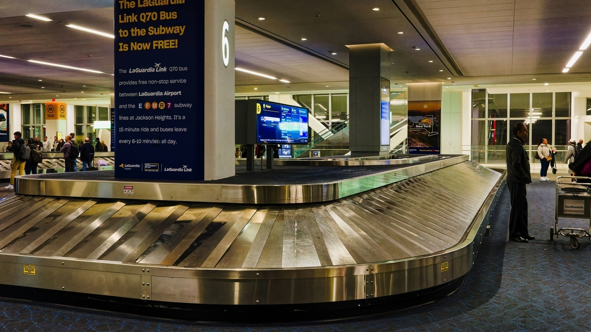 空港の到着ロビーにある手荷物受取所のターンテーブル。手前には大きな柱があり、バスの運行情報が書かれた青い看板が設置されています。奥の通路には、荷物を待つ数人の利用客の姿が見えます