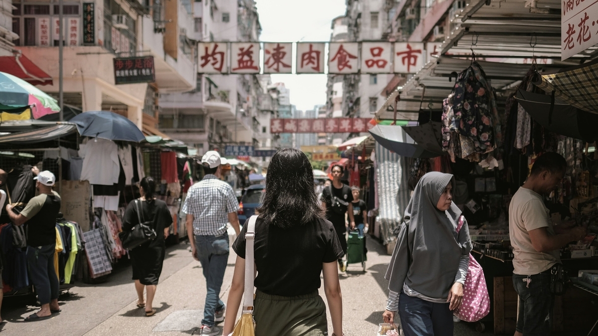 活気ある屋外市場の通りを歩く黒いTシャツ姿の女性の後ろ姿。通りには衣類を吊るした露店が並び、頭上には「均益凍肉食品市場」と書かれた看板が掲げられています。周囲には買い物客や歩行者が行き交っています