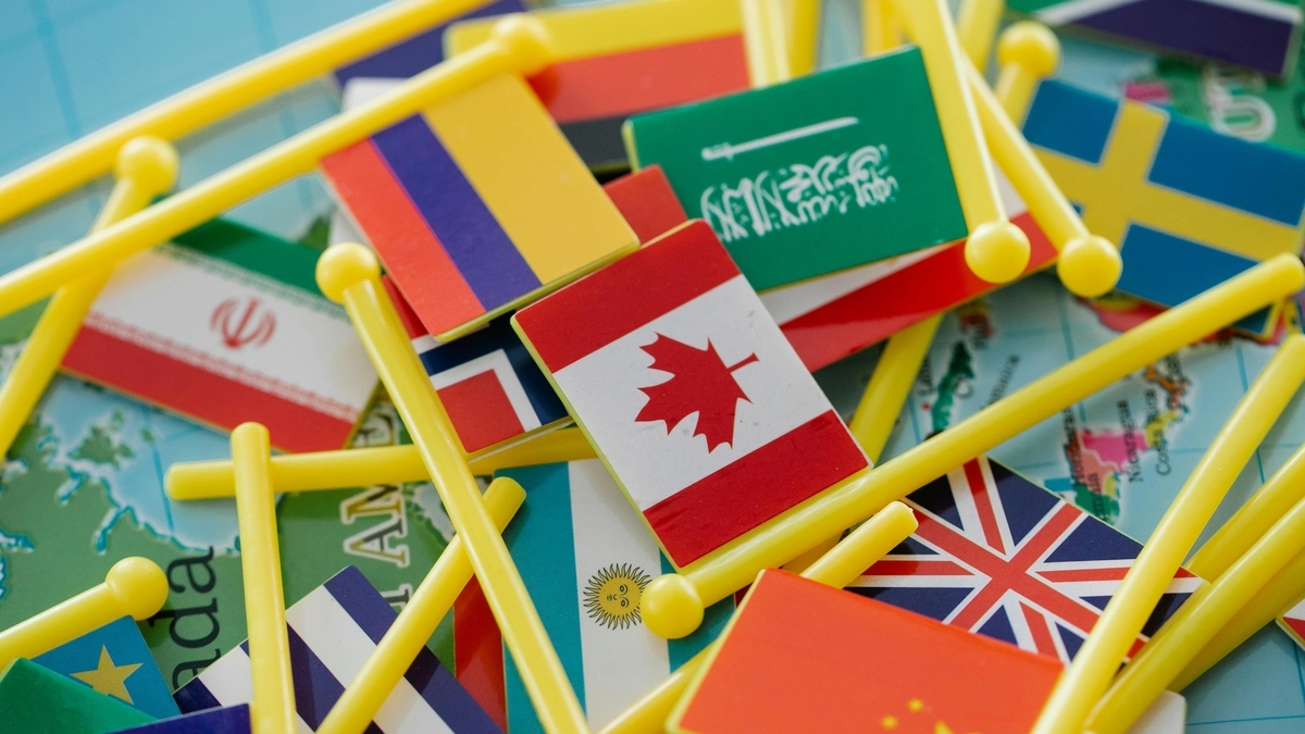 世界地図の上に散らばる、カナダ、イギリス、中国、サウジアラビアなど様々な国の小さな旗の模型。黄色いプラスチックの棒が付いた旗が重なり合い、その下には地図の地名や境界線が見えています