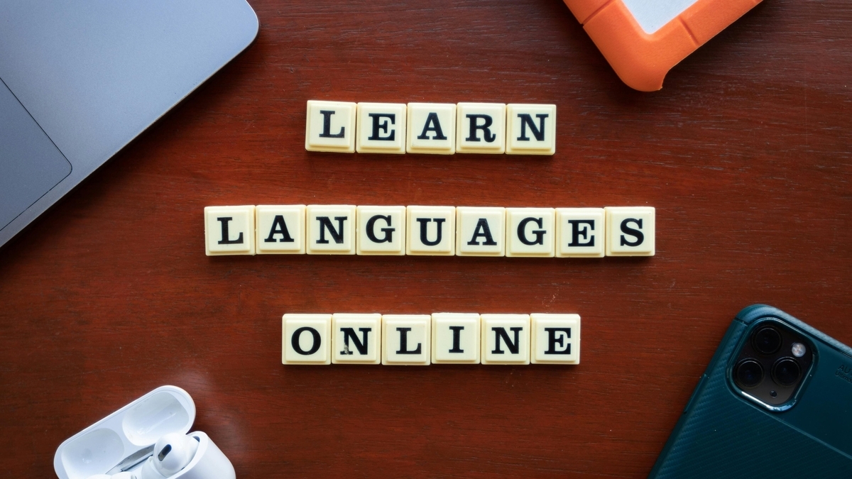 茶色の木製デスクの上に、「LEARN LANGUAGES ONLINE」と書かれた白いタイルの文字。周囲にはノートパソコン、スマートフォンのほか、ワイヤレスイヤホンやオレンジ色のケースが配置されています
