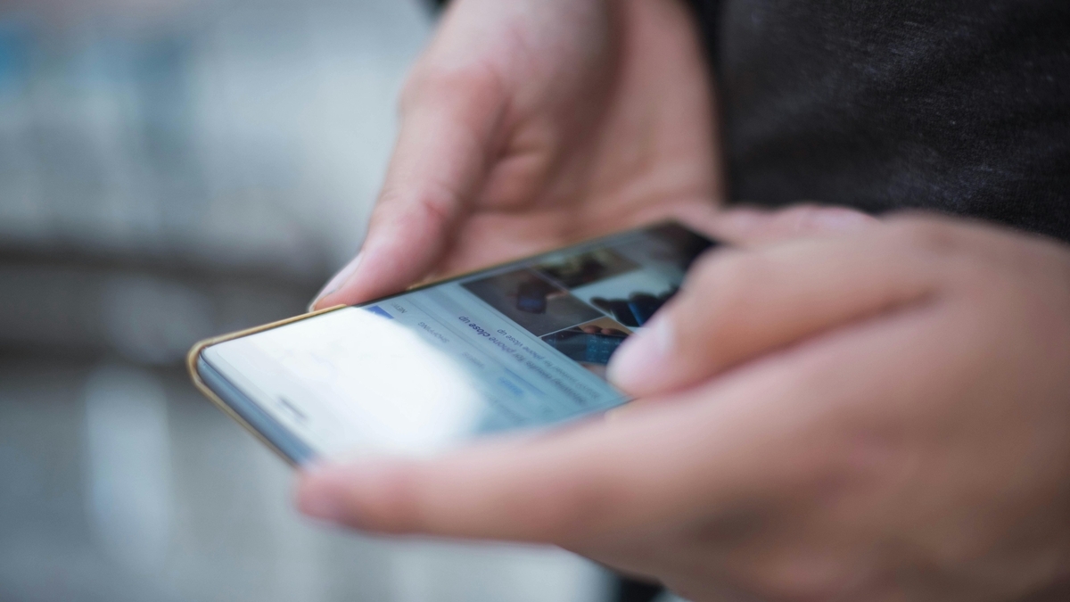両手でスマートフォンを持ち、画面を操作している人物の手元のアップ。スマートフォンの画面には画像が並ぶウェブサイトが表示されており、指先でスクロールしている様子が捉えられています。