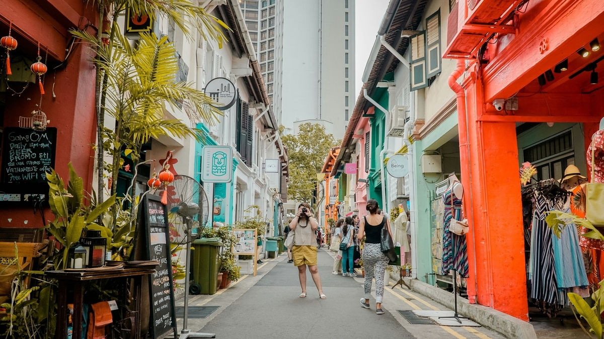 カラフルな建物が並ぶ狭い路地で、カメラを構える女性や歩く人々。店頭にはメニューボードや衣類が並び、上空には高層ビルがそびえ立つ、異国情緒漂う賑やかな通り