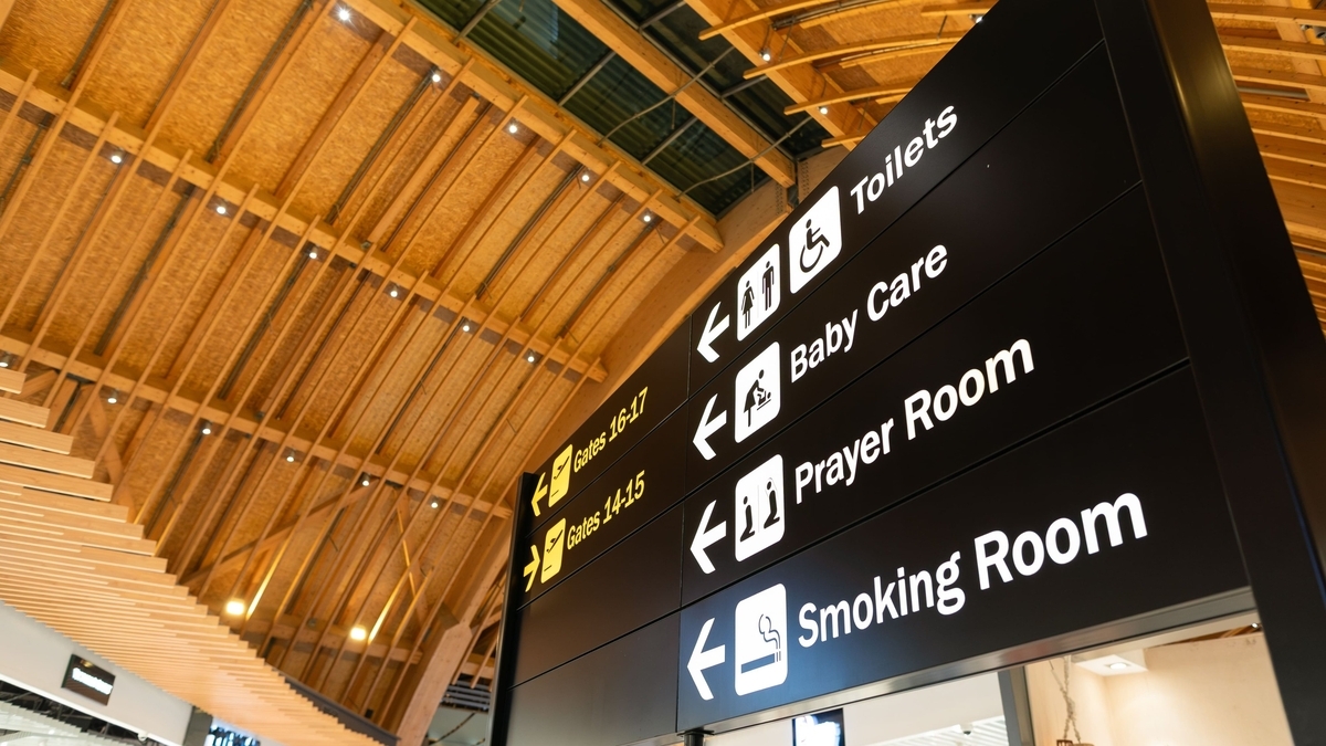空港内のような施設にある黒い案内看板。下からSmoking Room、Prayer Room、Baby Care、Toiletsと白文字で記され、それぞれにピクトグラムと左を指す矢印が添えられています