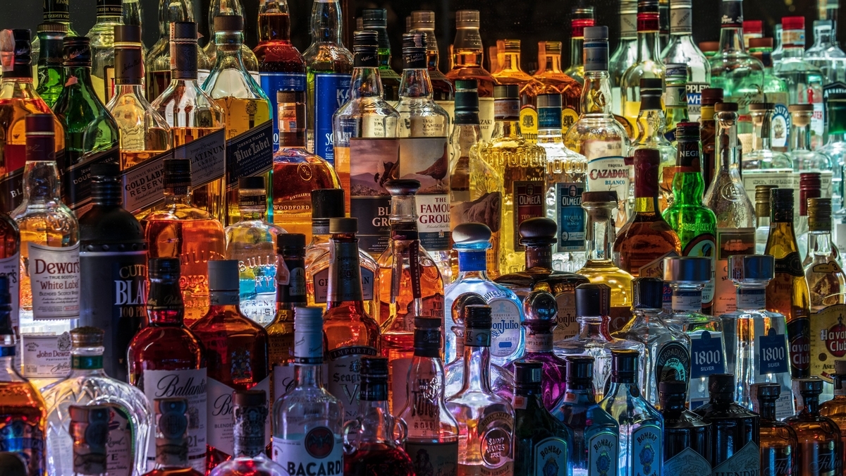 バックライトに照らされたバーの棚に、多種多様な洋酒のボトルが隙間なく並んでいる様子。Dewar'sやBallantine's、BACARDIなどのラベルが見え、琥珀色や透明、青色の瓶が鮮やかに輝いています