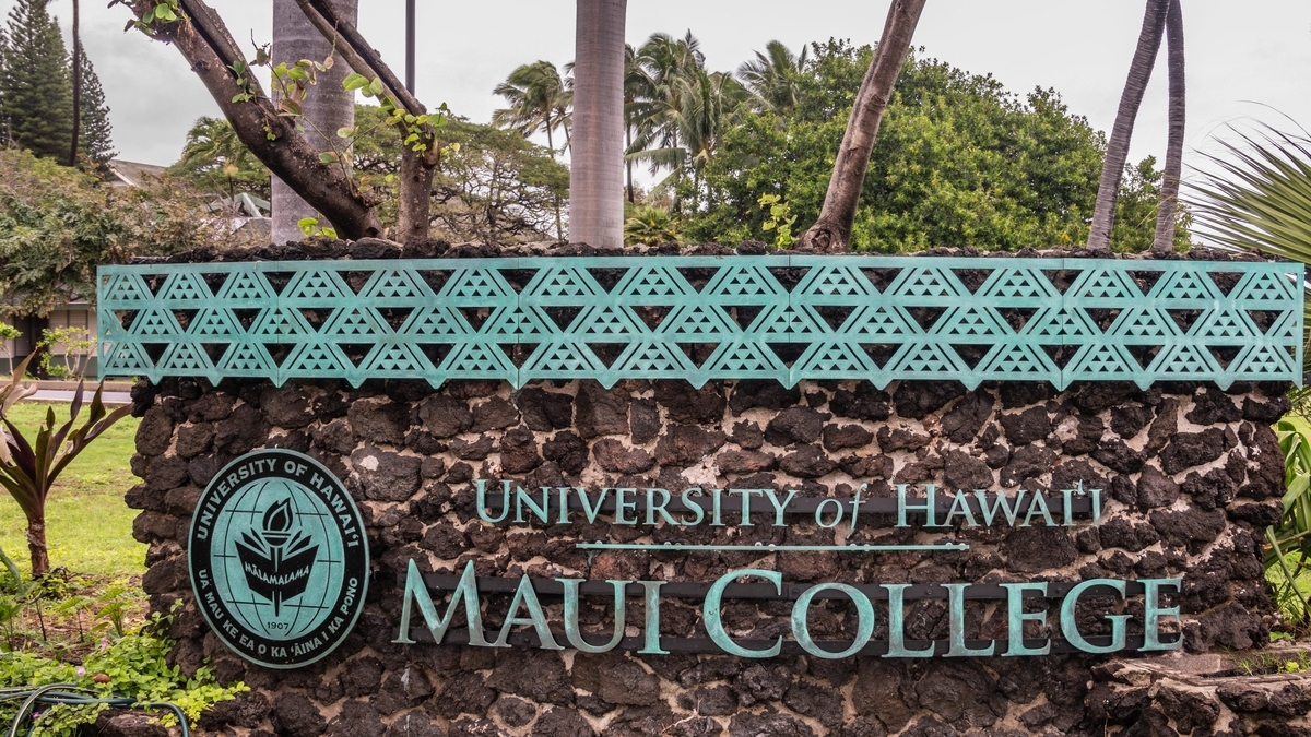 ゴツゴツとした石造りの壁に設置された「UNIVERSITY of HAWAI'I MAUI COLLEGE」の看板。左側には大学のロゴマークが配置され、上部には幾何学模様の透かし彫りがある青緑色の装飾が施されています