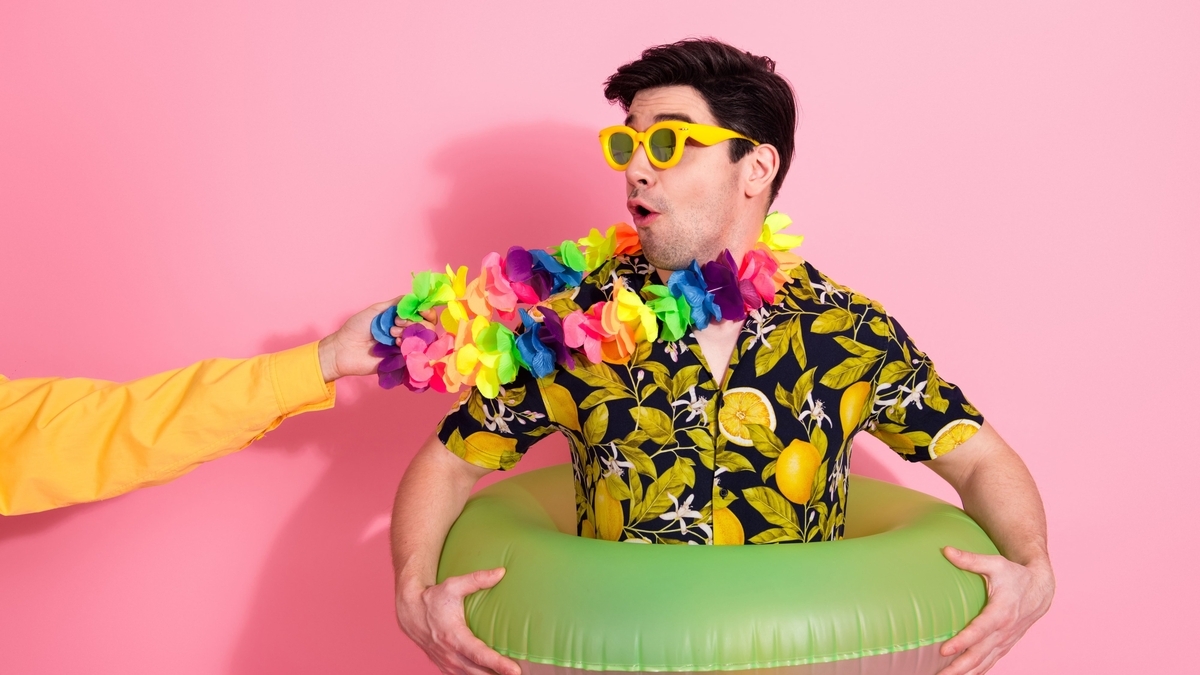 ピンク色の背景で、黄色のサングラスとレモン柄のシャツを着用した男性が、緑色の浮き輪を腰に通している姿。横から伸びてきた手にカラフルな花冠を首にかけられ、驚いたような表情で横を見ています。
