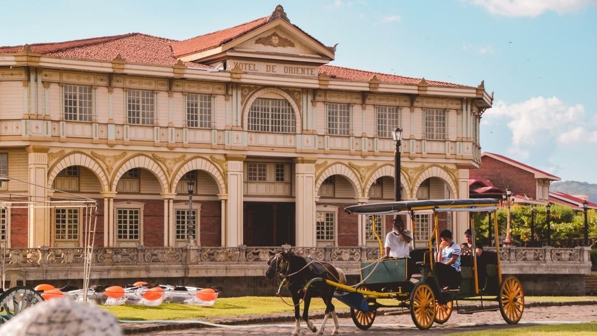 スペイン植民地時代の面影を残す、石造りの1階部分と木造の2階部分を持つ「HOTEL DE ORIENTE」の建物前。その手前の通りを、客を乗せた1頭立ての馬車が左から右へ進んでいる。