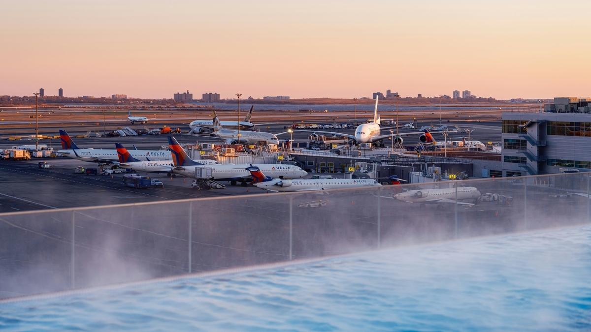 "夕暮れ時の空港に駐機するデルタ航空などの大型旅客機。手前には湯気が立ち上る屋外プールがあり、ガラス越しに滑走路や遠くの街並みを一望できます。オレンジ色に染まる空が機体や空港施設を照らしています