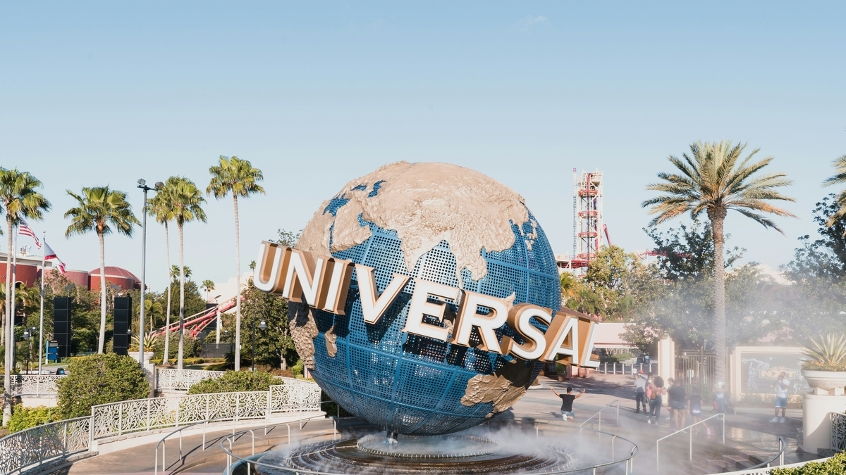 青空の下に立つ「UNIVERSAL」の文字が書かれた巨大な地球儀のモニュメント。周囲にはヤシの木が並び、霧状の噴水が上がっています。奥には赤いローラーコースターのレールや鉄塔が見える活気ある屋外の風景です