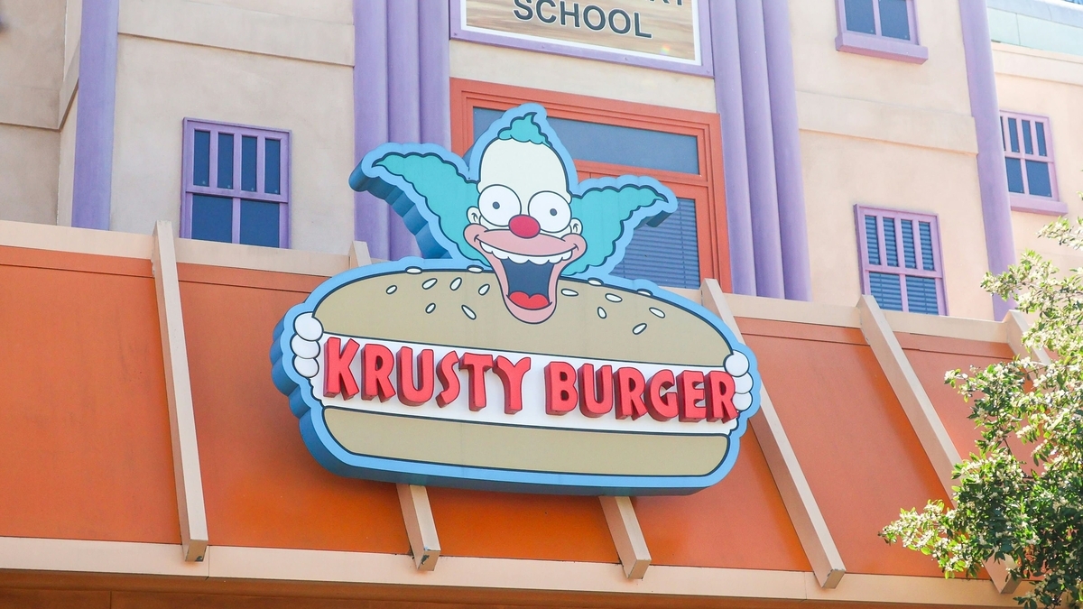 ピエロのキャラクターがハンバーガーを掲げた「KRUSTY BURGER」の看板。オレンジ色の屋根の上に設置されており、背後の建物は薄紫色の柱と窓が並ぶカートゥーン風のデザインで構成されています