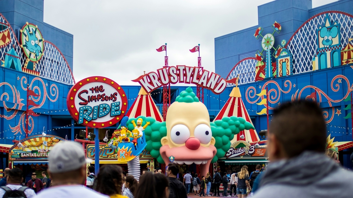 「KRUSTYLAND」と書かれたアーチの下に、ピエロのキャラクターの巨大な顔が設置されたアトラクションの入り口。左側には「THE SIMPSONS RIDE」の丸い看板があり、大勢の人々がカラフルな建物に向かって歩いています