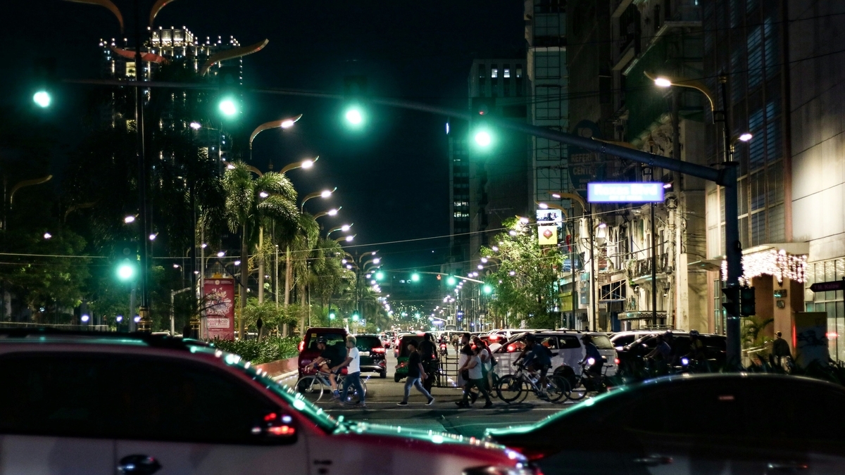 夜の街路樹がライトアップされた大通りを、多くの自動車や自転車が行き交う様子。奥には高いビルが立ち並び、緑色の街灯が等間隔に並んでいる