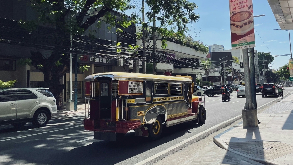 晴れた日の道路を走行する、黄色い屋根と装飾が特徴的な乗合バスのジープニー。街路樹の影が落ちる路肩には看板や電柱が立っている