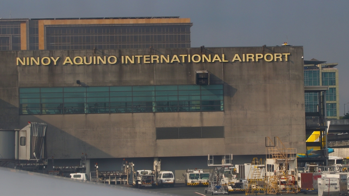 コンクリートの壁面に「NINOY AQUINO INTERNATIONAL AIRPORT」と書かれた空港の外観。建物の前には、タラップ車や牽引車などの地上作業車両が多数並んで停車している。