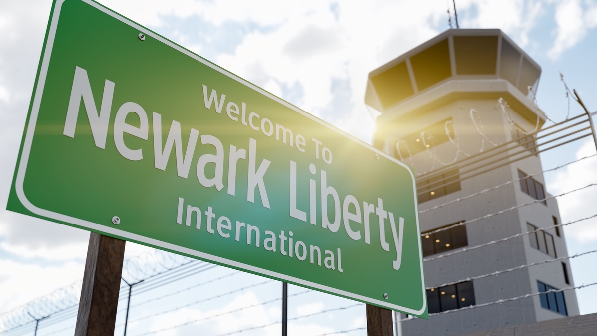 緑色の看板に白文字で「Welcome To Newark Liberty International」と記されています。背景にはフェンス越しに空港の管制塔がそびえ立ち、上空は雲が広がる空模様です。
