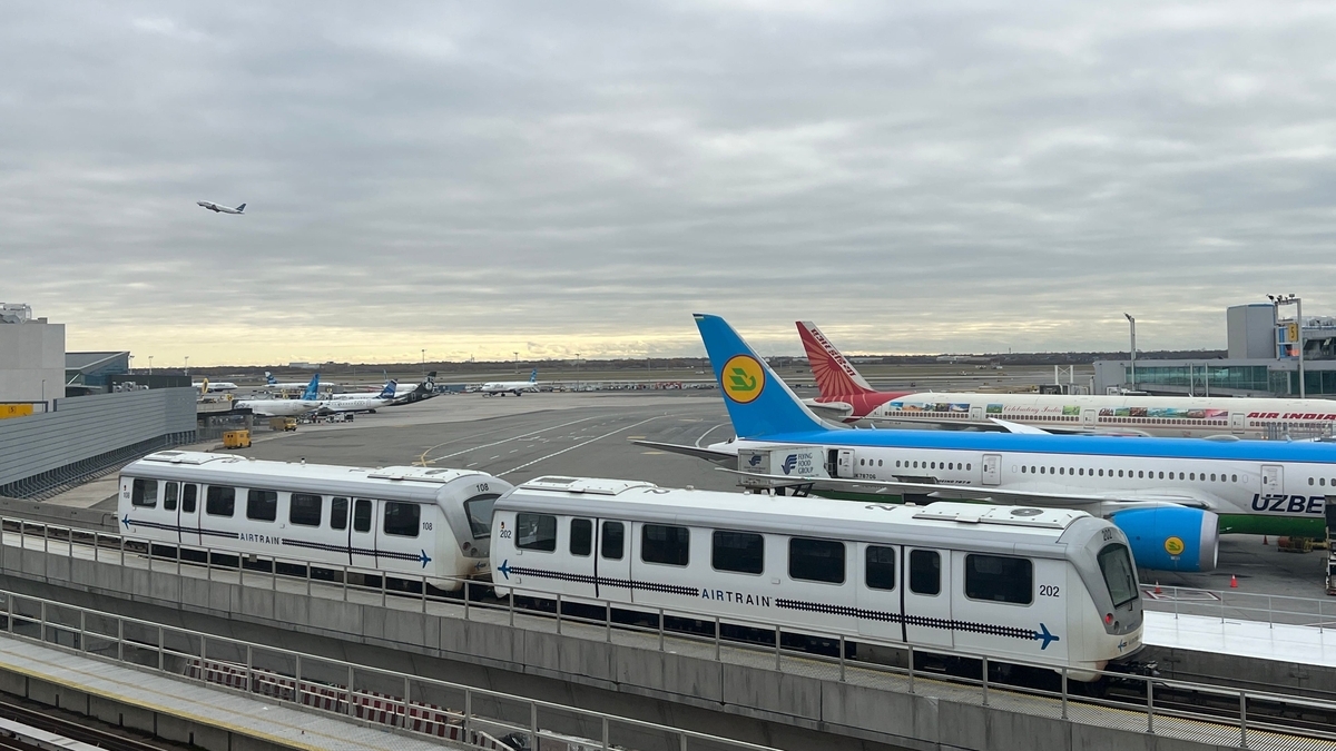 高架の上を白いAIRTRAINが走行しており、その背景には広大な空港の敷地が見えます。駐機している複数の航空機のほか、曇り空の下を離陸して遠ざかっていく機体も写り込んでいます。