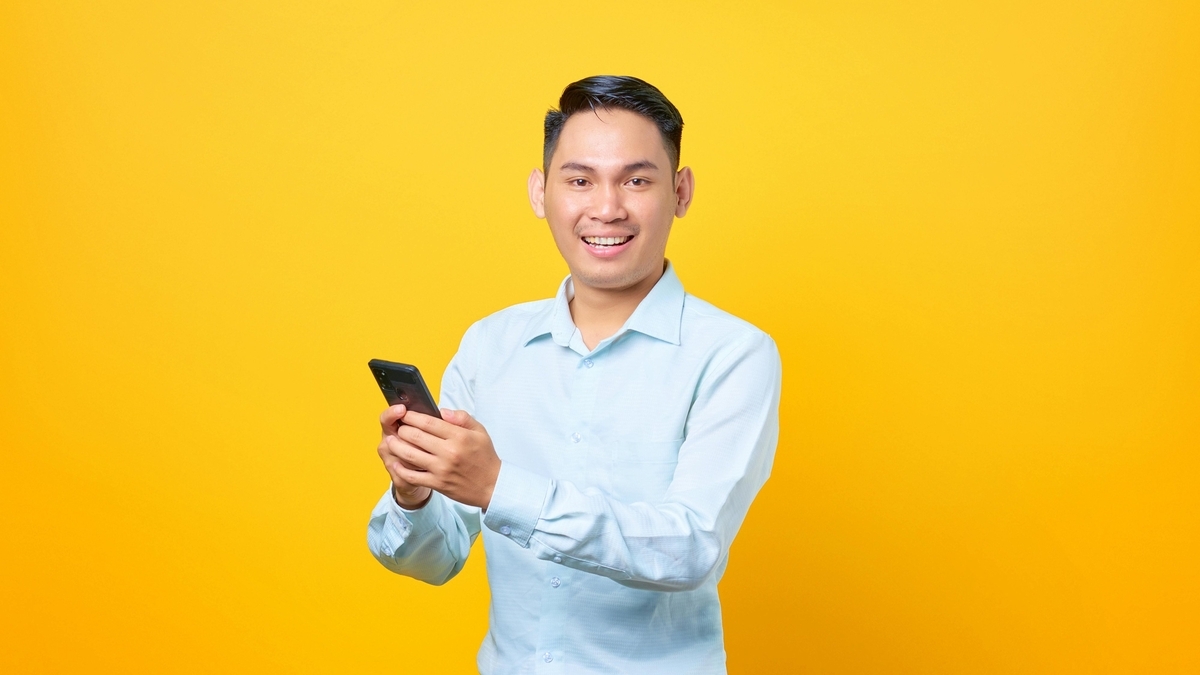 鮮やかな黄色の背景の前で、水色のシャツを着た若い男性がスマートフォンを両手で持ち、笑顔で正面を向いている。短髪で清潔感があり、デバイスを操作している瞬間を捉えた写真