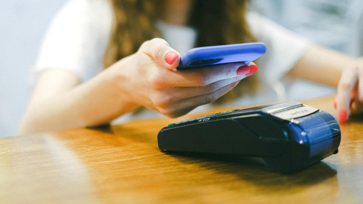 赤いネイルを施した女性の手が、青いケースに入ったスマートフォンをキャッシュレス決済端末にかざしている。木目のテーブルの上で、非接触決済を利用して支払う手元のアップ