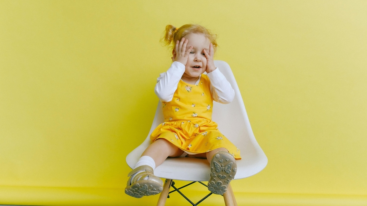黄色の背景の前で、花柄の黄色いワンピースを着た幼い少女が白い椅子に座っている。両手で顔を覆いながら、少し口を開けて楽しそうに笑っている表情を見せるポートレート。